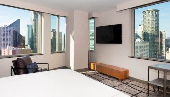 A modern bedroom with large windows showcasing a cityscape, a wall-mounted TV, an armchair, and a wooden bench.