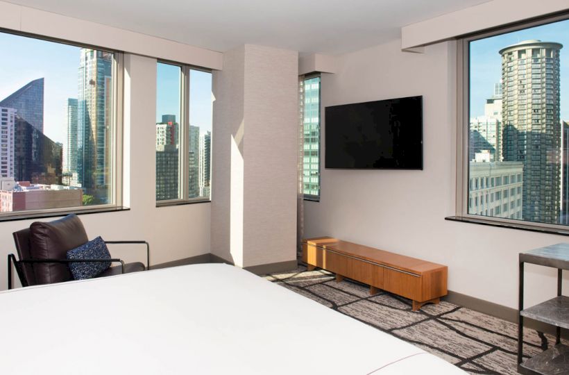 A modern bedroom with large windows showcasing a cityscape, a wall-mounted TV, an armchair, and a wooden bench.