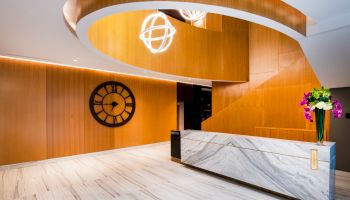 A modern lobby featuring a marble reception desk, a large wall clock, contemporary lighting fixtures, and a vase with colorful flowers.