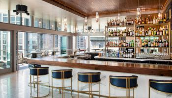 This image shows a modern bar with a wooden ceiling, a well-stocked bar, pendant lights, and high stools. Large windows provide a city view.