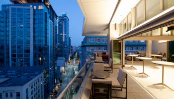 A modern urban balcony at dusk with city skyscrapers in the background; the area features seating and tables, and overlooks a busy street.