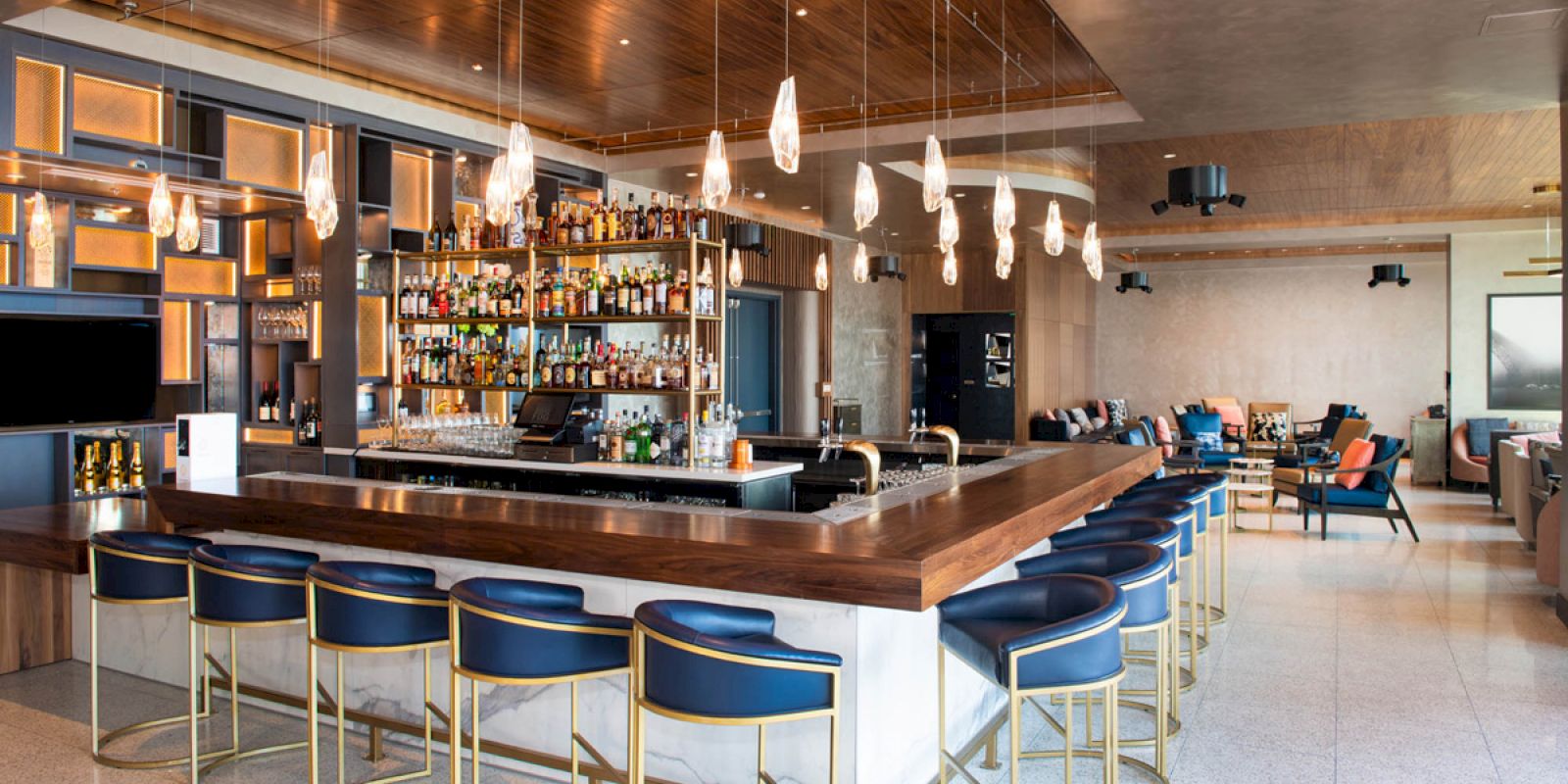 The image shows a modern, elegant bar with a wooden counter, blue chairs, hanging lights, and shelves stocked with various bottles.