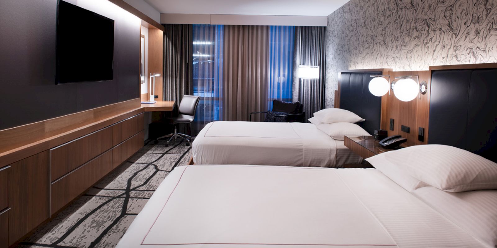 The image shows a modern hotel room with two neatly made beds, a workspace with a chair and desk, a flat-screen TV, and a large window with curtains.