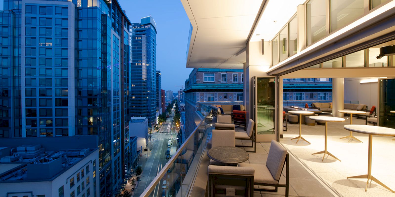 This image shows a modern cityscape with tall buildings at dusk, viewed from a balcony with lounge furniture and glass railings.