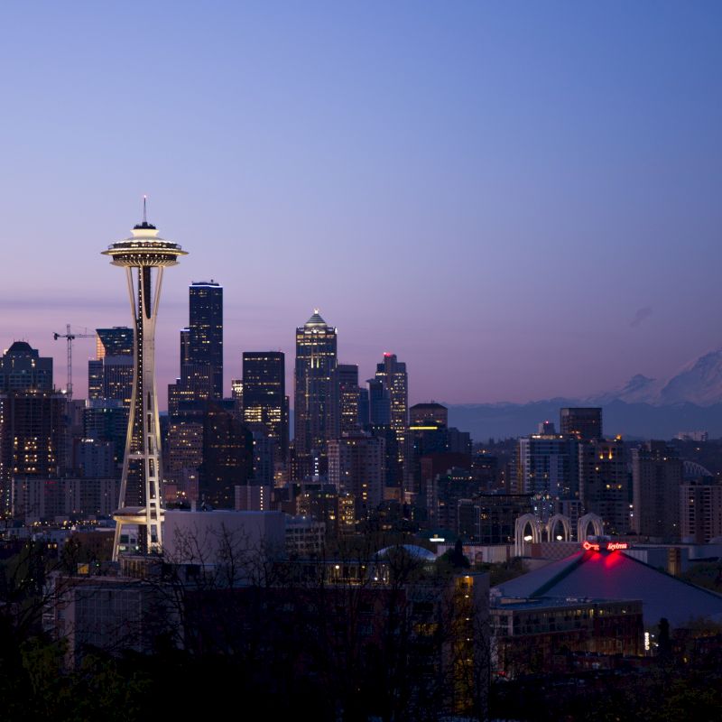 This image shows the Seattle skyline at dusk, featuring the Space Needle and Mount Rainier in the background, with city lights beginning to illuminate.