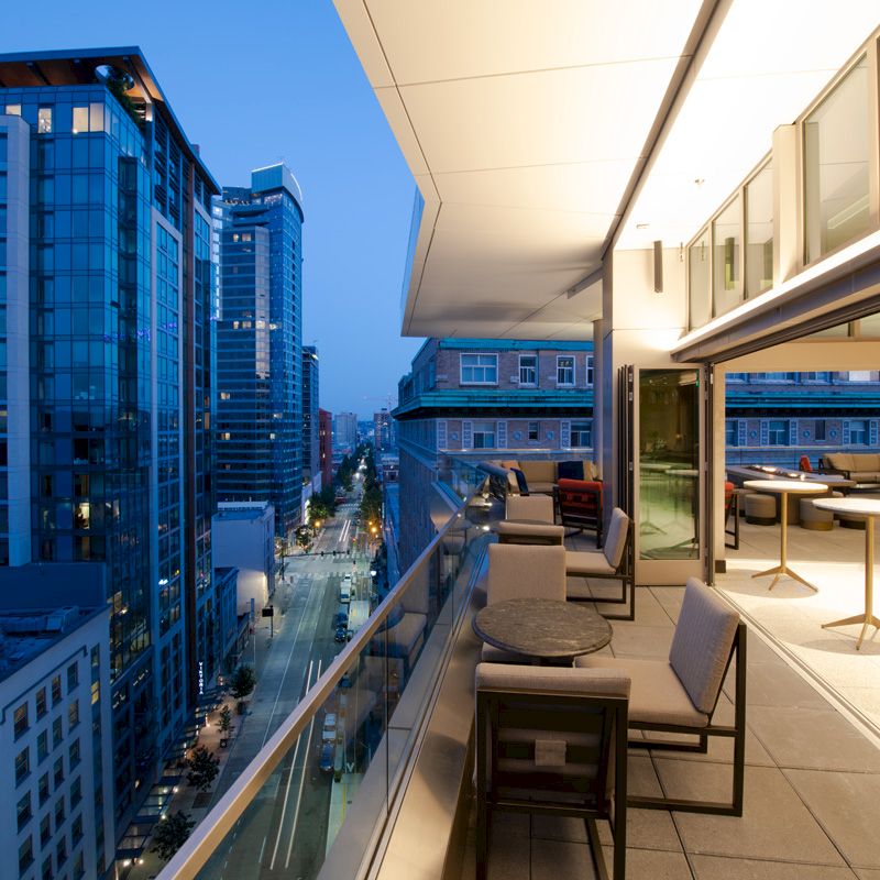 A modern balcony overlooks a cityscape at dusk, with stylish seating and tables. High-rise buildings line the street below, lights are on.