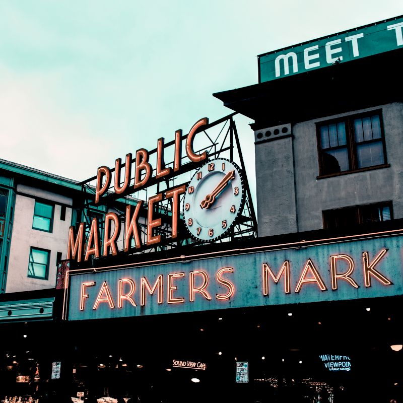 A neon signage says "PUBLIC MARKET" and "FARMERS MARKET" with a clock in the center, next to buildings with signs that include "MARKET HOTEL."