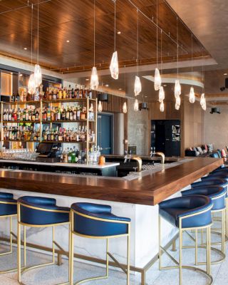 A modern bar with a wooden countertop, blue chairs, hanging lights, and well-organized shelves of bottled drinks, with cozy seating in the background.