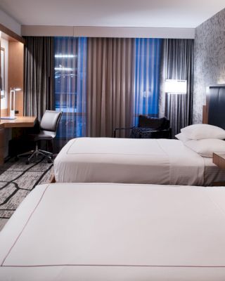 A modern hotel room with two single beds, a wall-mounted TV, a desk, and large windows with curtains. The room features contemporary decor.
