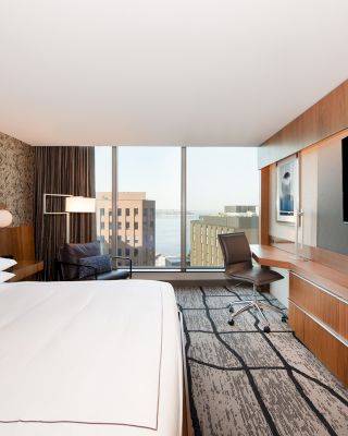 A modern hotel room with a large bed, TV on a wooden panel, desk, chair, and a window overlooking buildings and possibly a body of water outside.