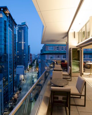 A modern cityscape view from a high-rise balcony at dusk, featuring outdoor seating and tables, with tall buildings and a street below.