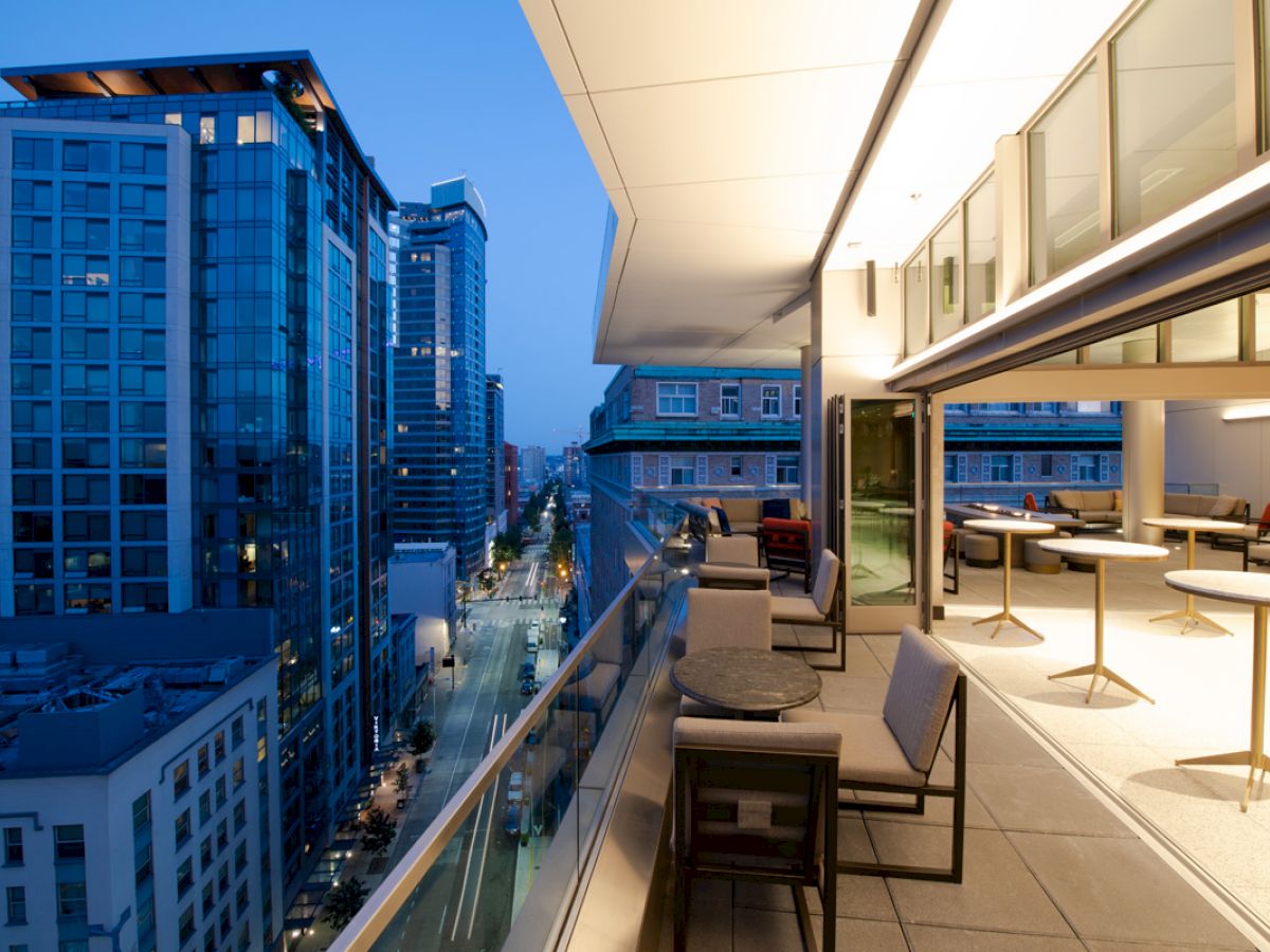 A modern urban balcony at dusk with glass railings, seating, and buildings lining the street below, illuminated by city lights and building interiors.
