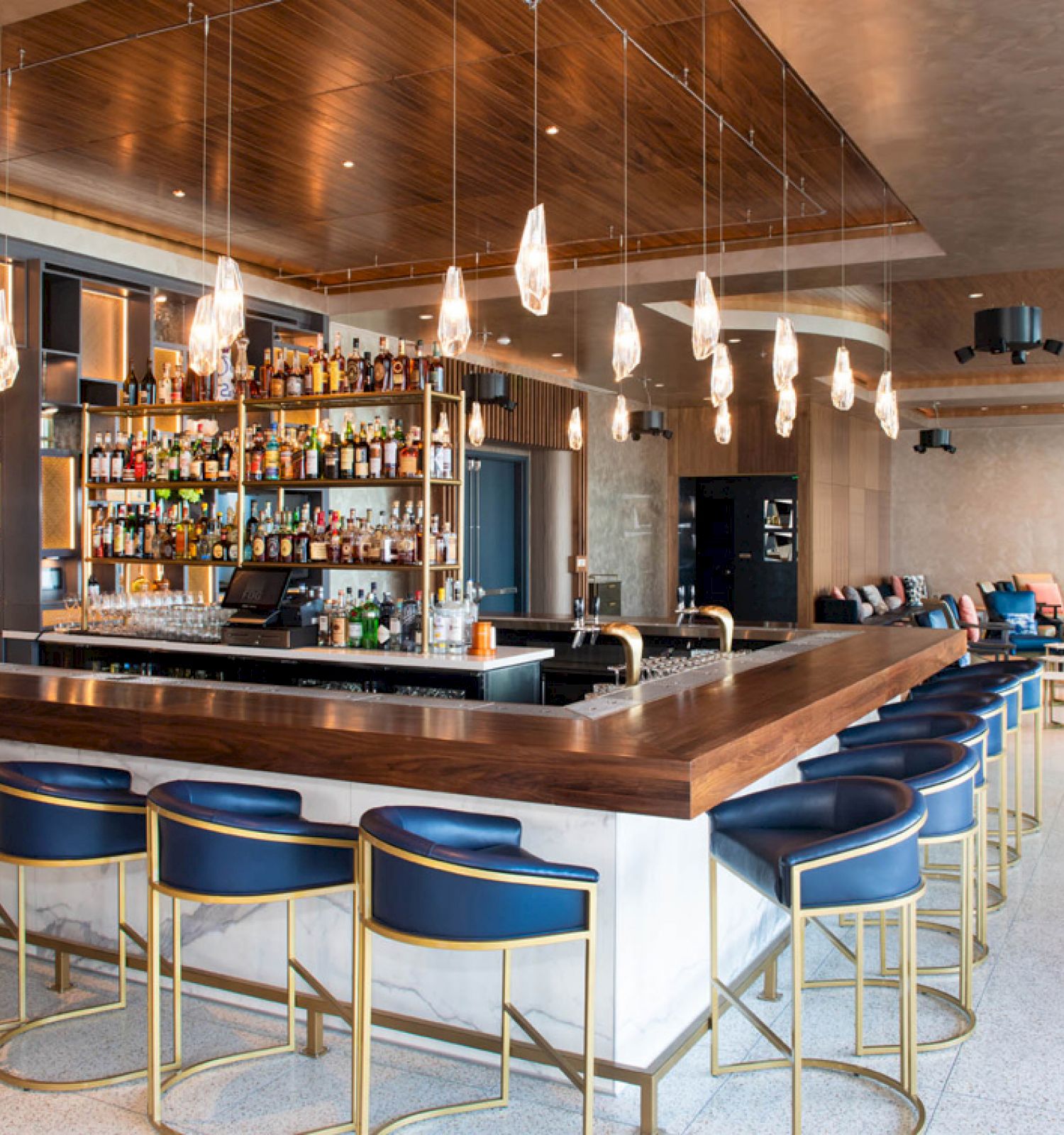The image shows a modern bar with a U-shaped counter, blue stools, a fully stocked bar, hanging lights, and a seating area in the background.