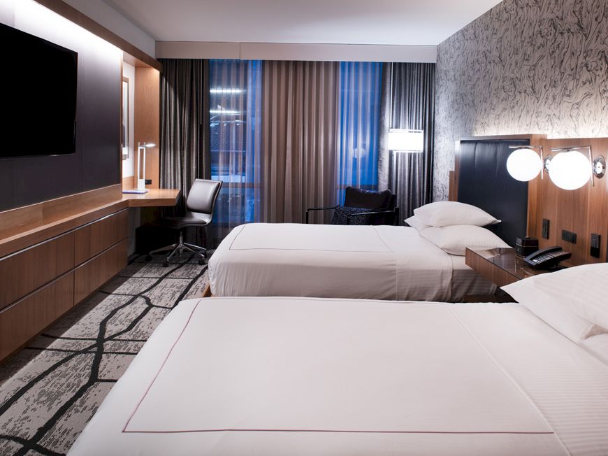 A modern hotel room with two beds, a wall-mounted TV, a desk, and a chair by a window with curtains.