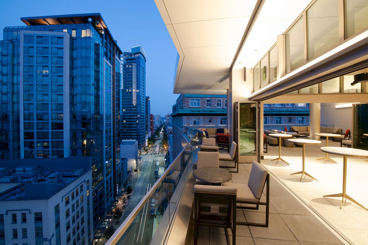 A modern balcony with seating and tables overlooks an urban cityscape at dusk, highlighted by tall buildings and street lights.