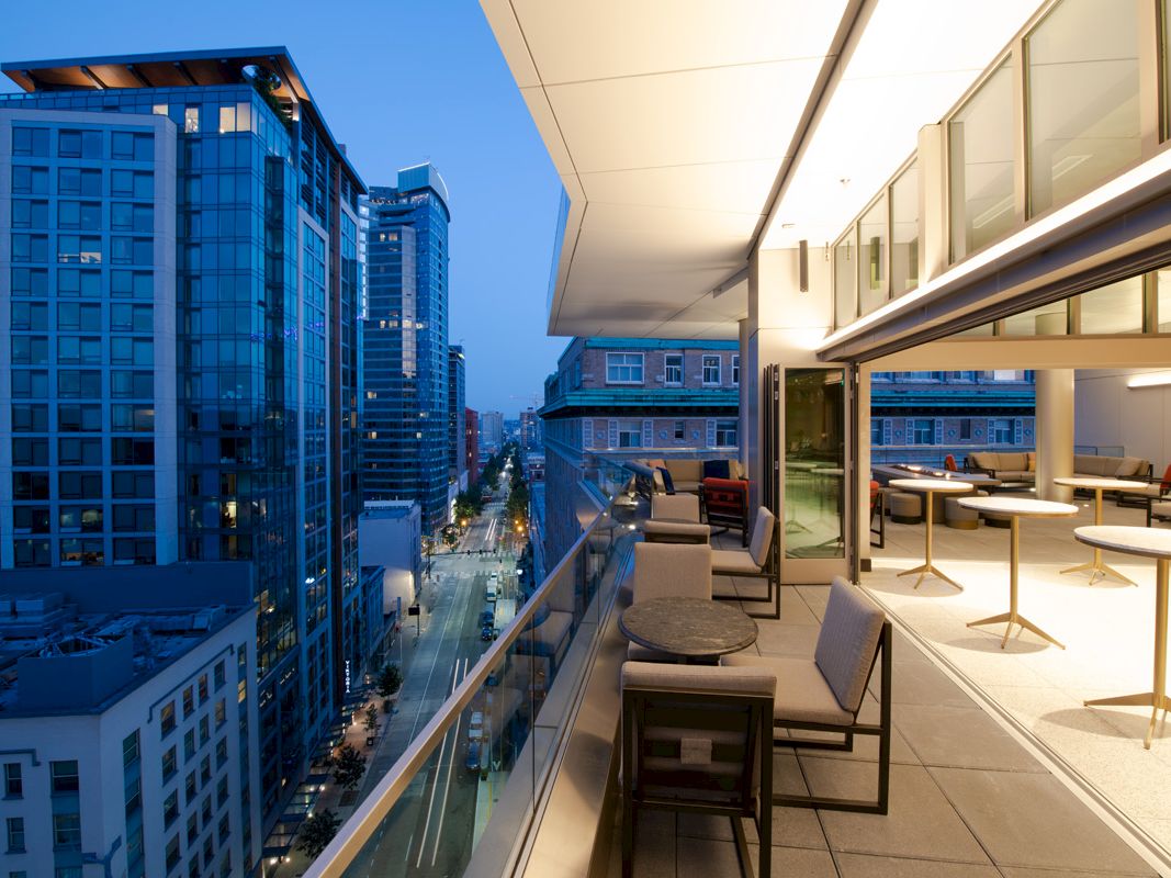 A modern balcony with seating and tables overlooks an urban cityscape at dusk, highlighted by tall buildings and street lights.