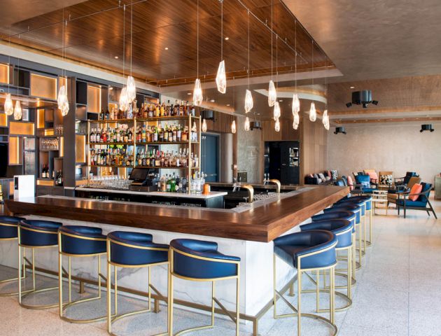 A modern, stylish bar with blue chairs, a well-stocked liquor shelf, hanging lights, and a spacious seating area in the background.