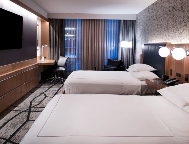 A modern hotel room with two neatly made beds, a flat-screen TV, a work desk, and stylish decor featuring a mix of wood and muted tones.
