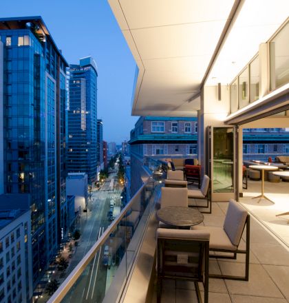 The image shows a modern cityscape at dusk with tall buildings, viewed from a balcony featuring tables, chairs, and seating areas.