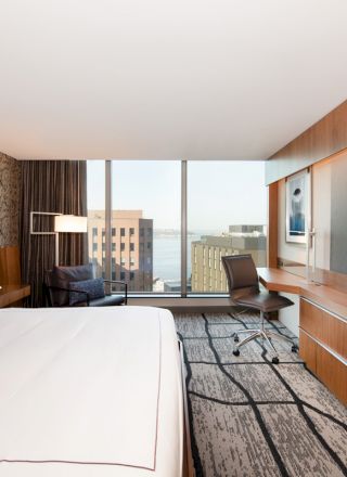 A modern hotel room features a large bed, flat-screen TV, desk with chair, and large windows with a city view.