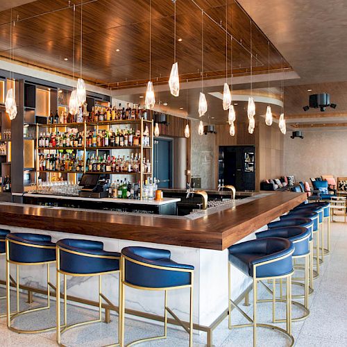 The image shows a modern bar with high stools, a well-stocked liquor shelf, hanging lights, and a seating area in the background.