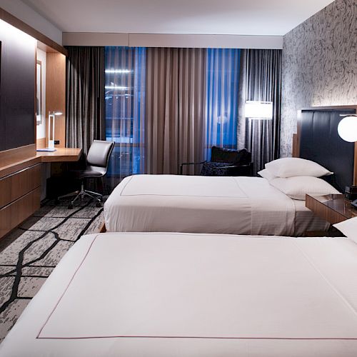 The image shows a modern hotel room with two neatly made beds, a desk with a chair, a flat-screen TV, and stylish decor and lighting.