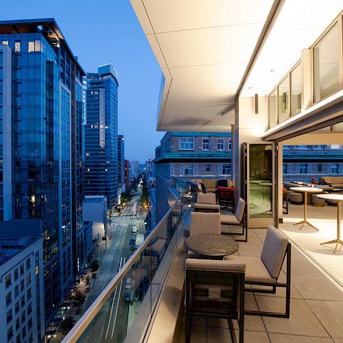 A modern urban balcony at dusk, offering views of city buildings and streets. The balcony has tables, chairs, and large windows.