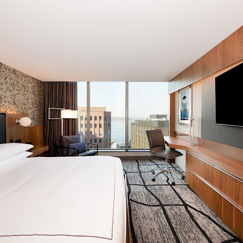 A modern hotel room with a large bed, a desk, a chair, a flat-screen TV, and a view of city buildings through a large window.