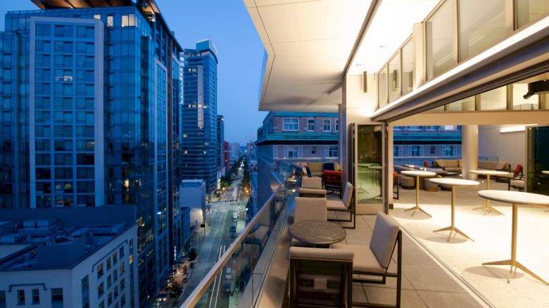 A modern balcony with tables and chairs overlooks an urban street surrounded by tall buildings, illuminated during twilight hours.
