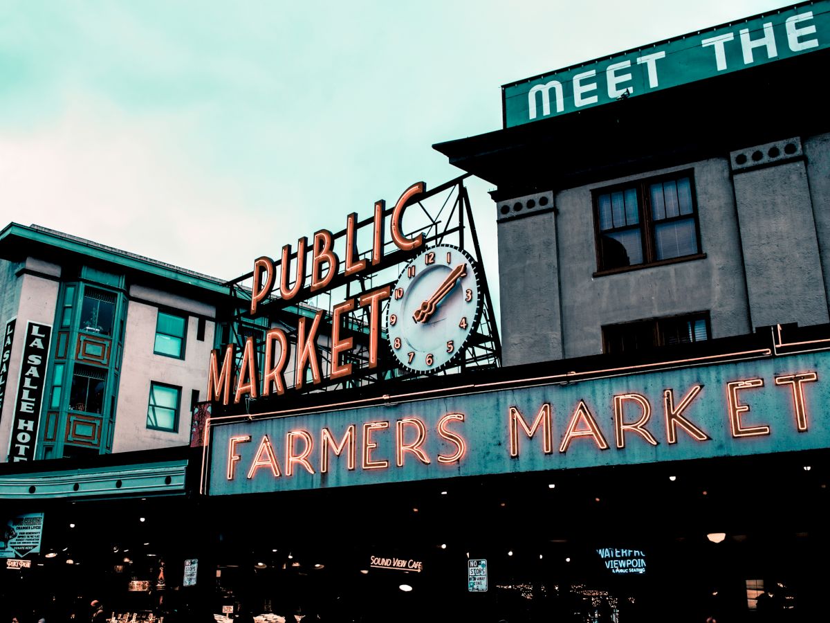 The image shows a public market with a neon "Farmers Market" sign and a building displaying "Meet the Producers" in large letters.