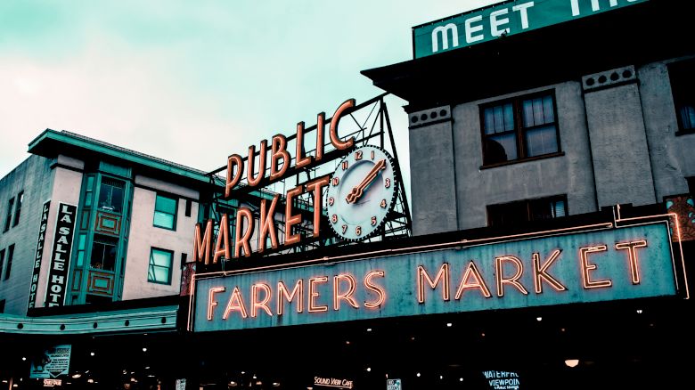 The image shows a public market with a neon "Farmers Market" sign and a building displaying "Meet the Producers" in large letters.