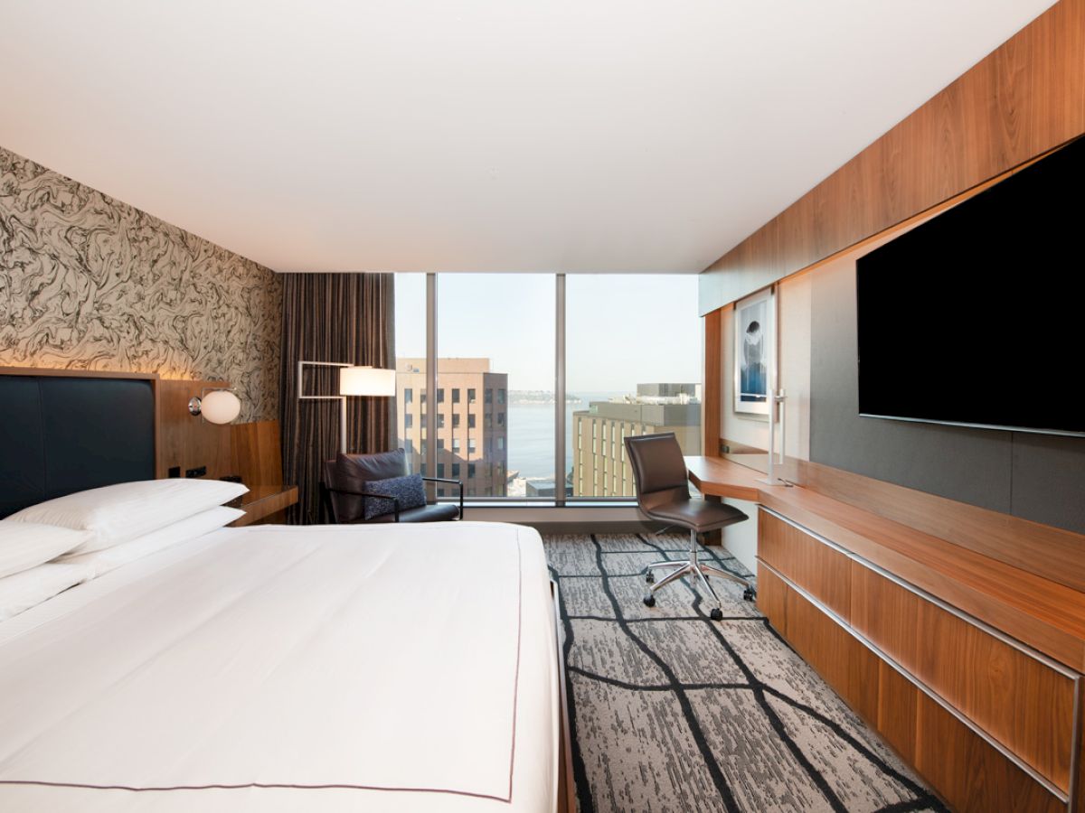 A modern hotel room with a large bed, a wall-mounted TV, a desk with a chair, a stylish chair by the window, and cityscape views outside.