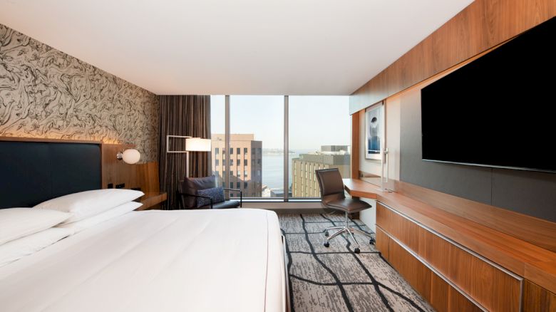 A modern hotel room with a large bed, a wall-mounted TV, a desk with a chair, a stylish chair by the window, and cityscape views outside.