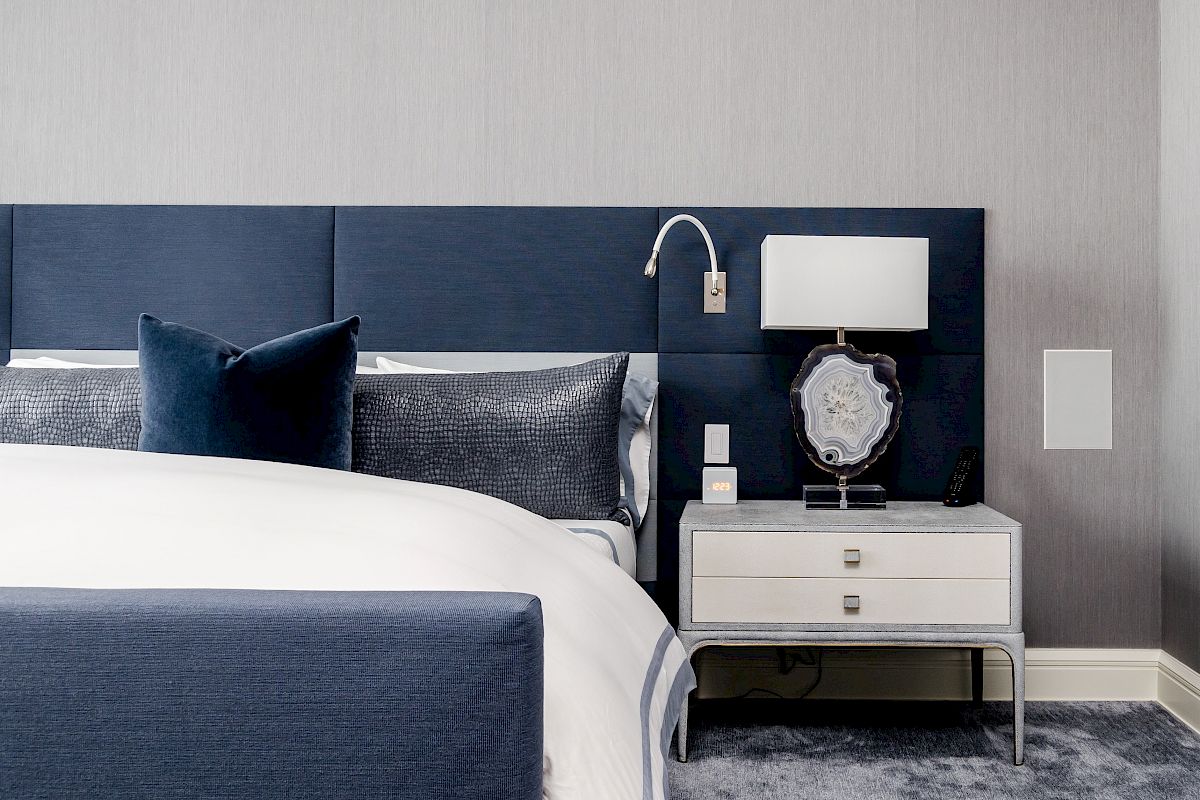 This image shows a modern bedroom with a navy blue headboard, white bedding, and a nightstand featuring a lamp and decorative items, ending the sentence.