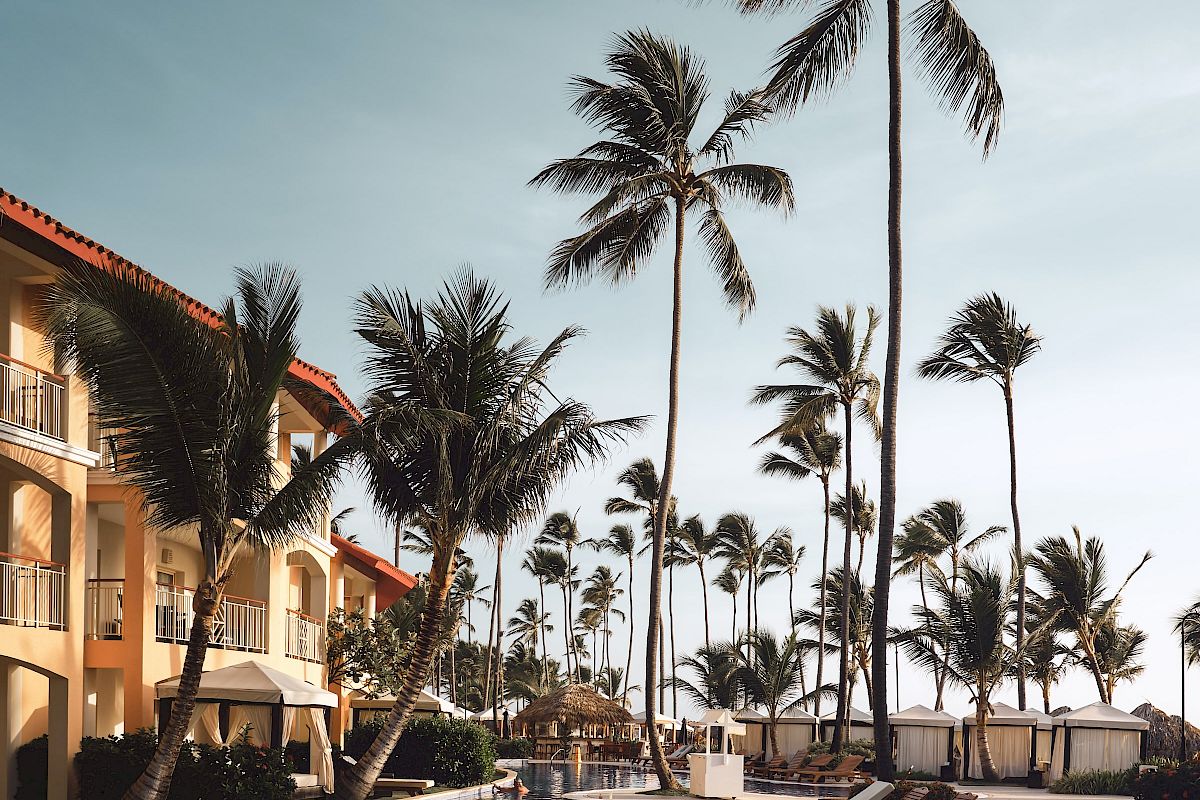 A luxurious poolside area lined with lounge chairs and tall palm trees next to a multi-story resort building under a clear sky.
