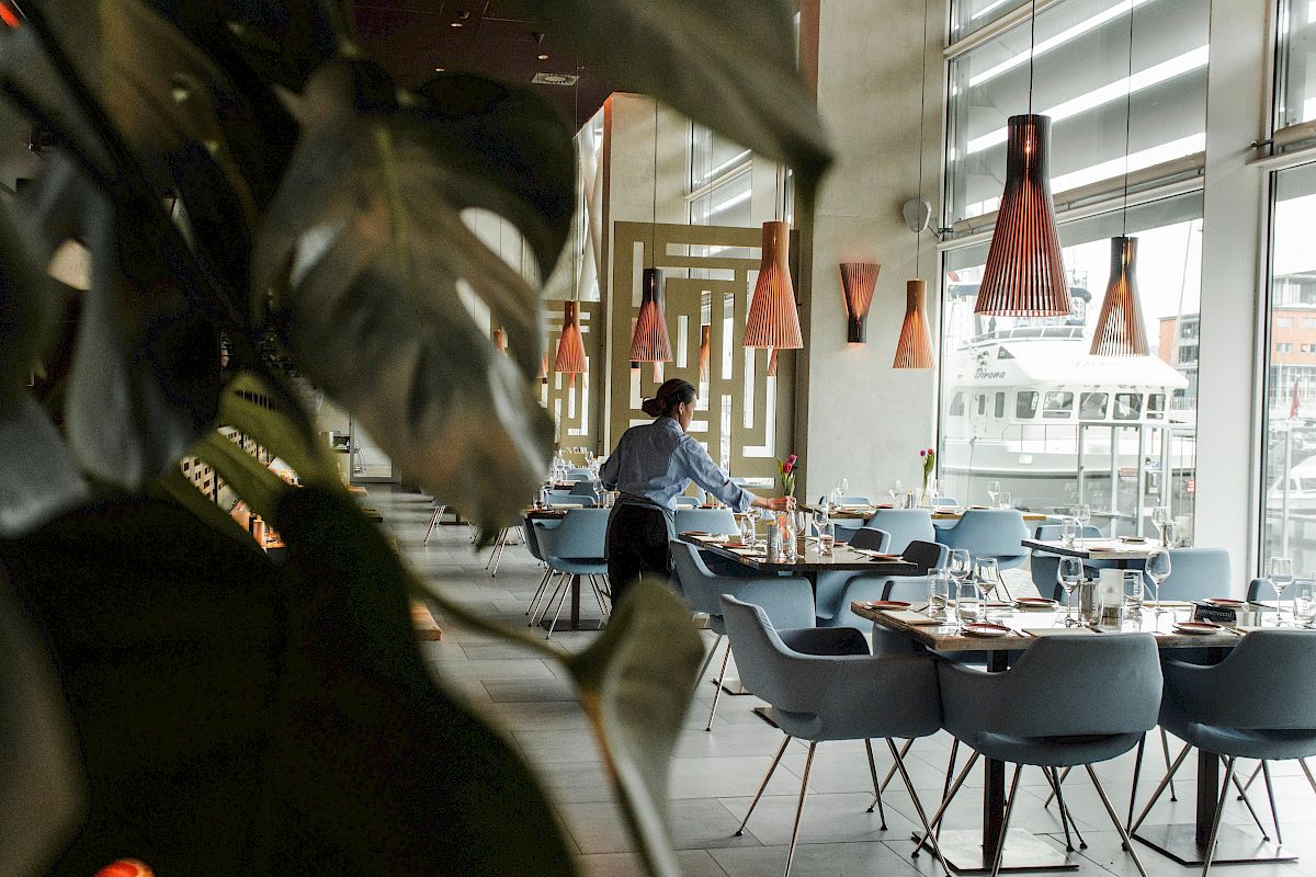 A person is setting tables in a stylish, modern restaurant with large windows, pendant lights, and blue chairs.