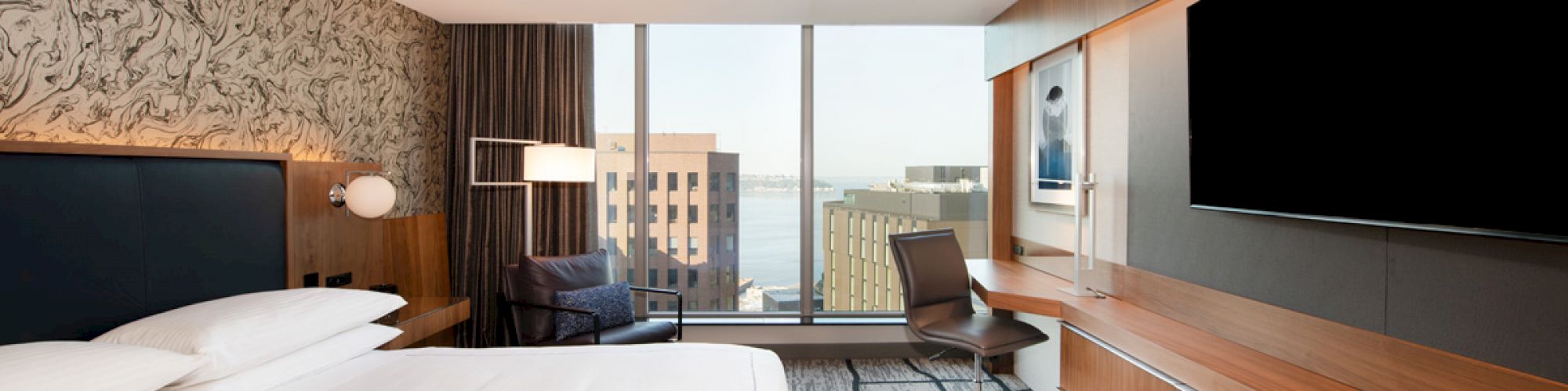 A modern hotel room featuring a large bed, a desk with a chair, a flat-screen TV, large windows with a city view, and stylish decor.