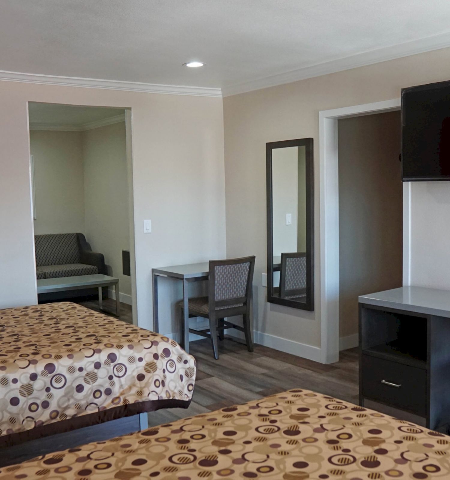 A hotel room with two beds, a desk with a chair, a wall-mounted TV, a mirror, and a small seating area in the corner.