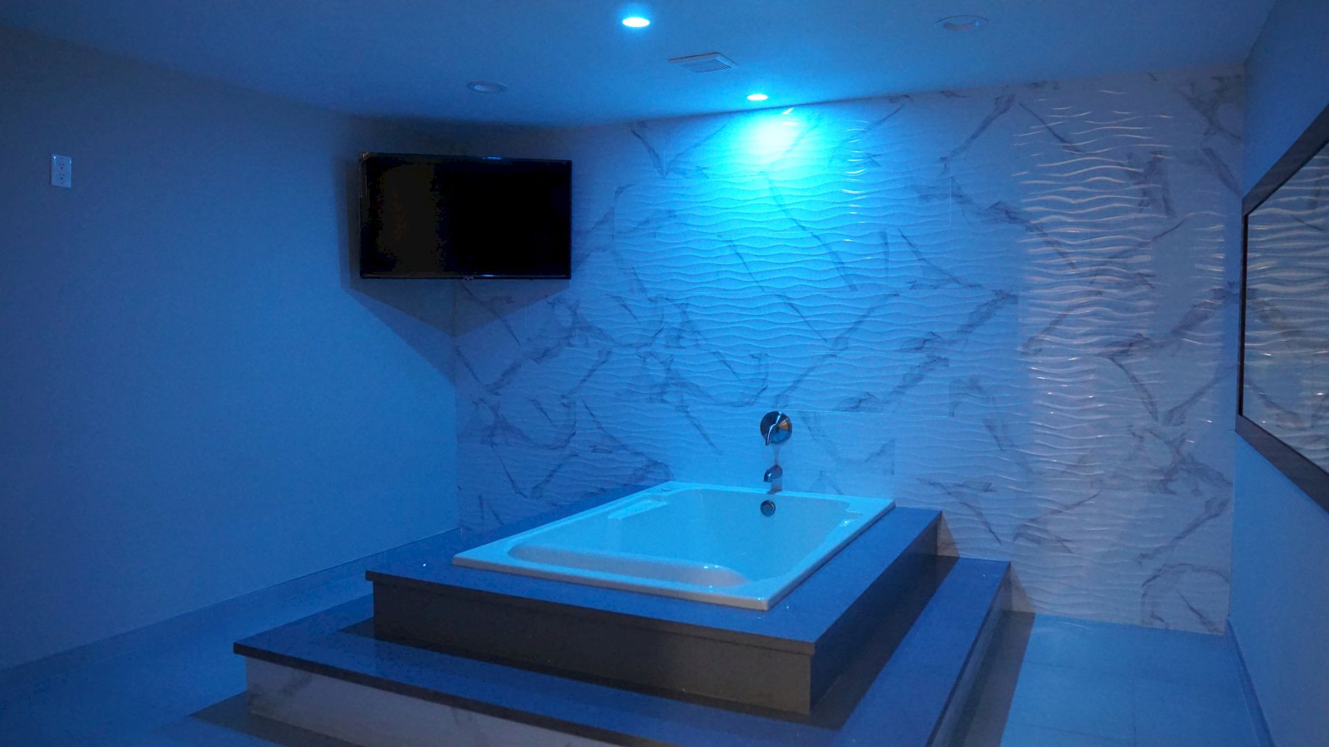A modern bathroom with a sunken tub, accented blue lighting, a wall-mounted TV, and a large textured wall.