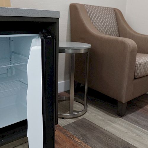 The image shows an open mini-fridge next to a small side table and an armchair in a room with wooden flooring and a large mirror.