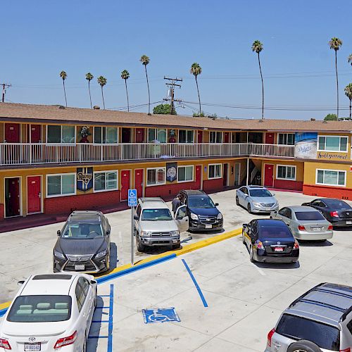 The image shows a motel courtyard with parked cars and a second-floor walkway, plus palm trees and a clear blue sky in the background.